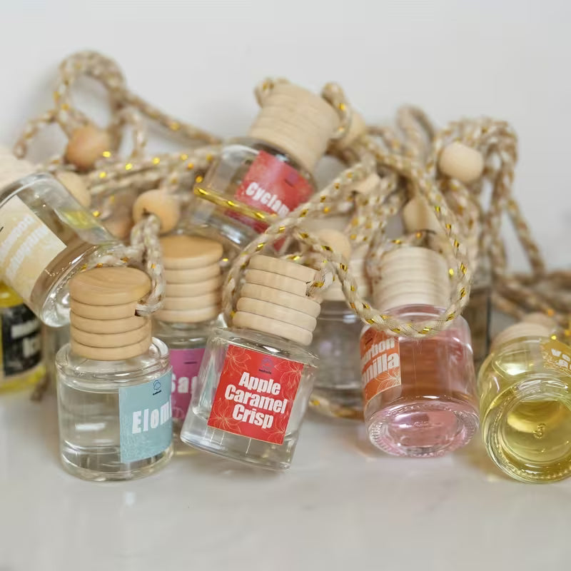 A group of small glass car air freshener  bottles with light wood caps and braided cord necks, arranged on a white surface. Visible labels include Elom, Apple Caramel Crisp, and Cinnamon.