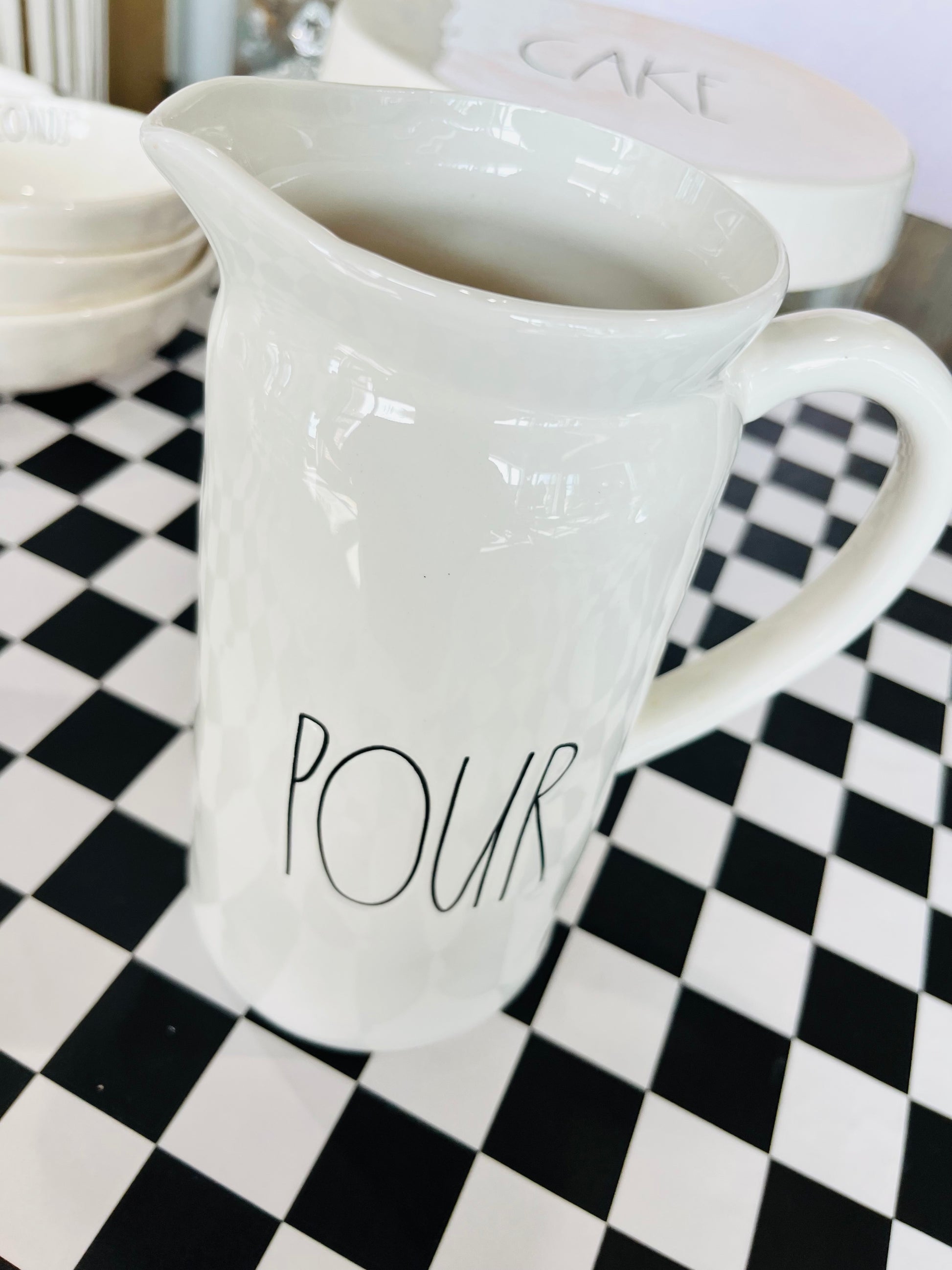 White ceramic pitcher with 'POUR' text on a black and white checkered surface