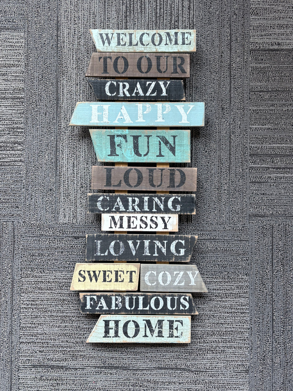 A vertical stack of rustic wooden sign slats with hand-painted distressed letters spelling out 'WELCOME TO OUR CRAZY HAPPY FUN LOUD CARING MESSY LOVING SWEET COZY FABULOUS HOME' against a textured gray wall.