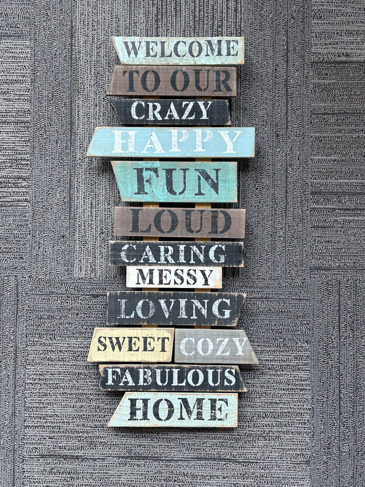 A vertical stack of rustic wooden sign slats with hand-painted distressed letters spelling out 'WELCOME TO OUR CRAZY HAPPY FUN LOUD CARING MESSY LOVING SWEET COZY FABULOUS HOME' against a textured gray wall.
