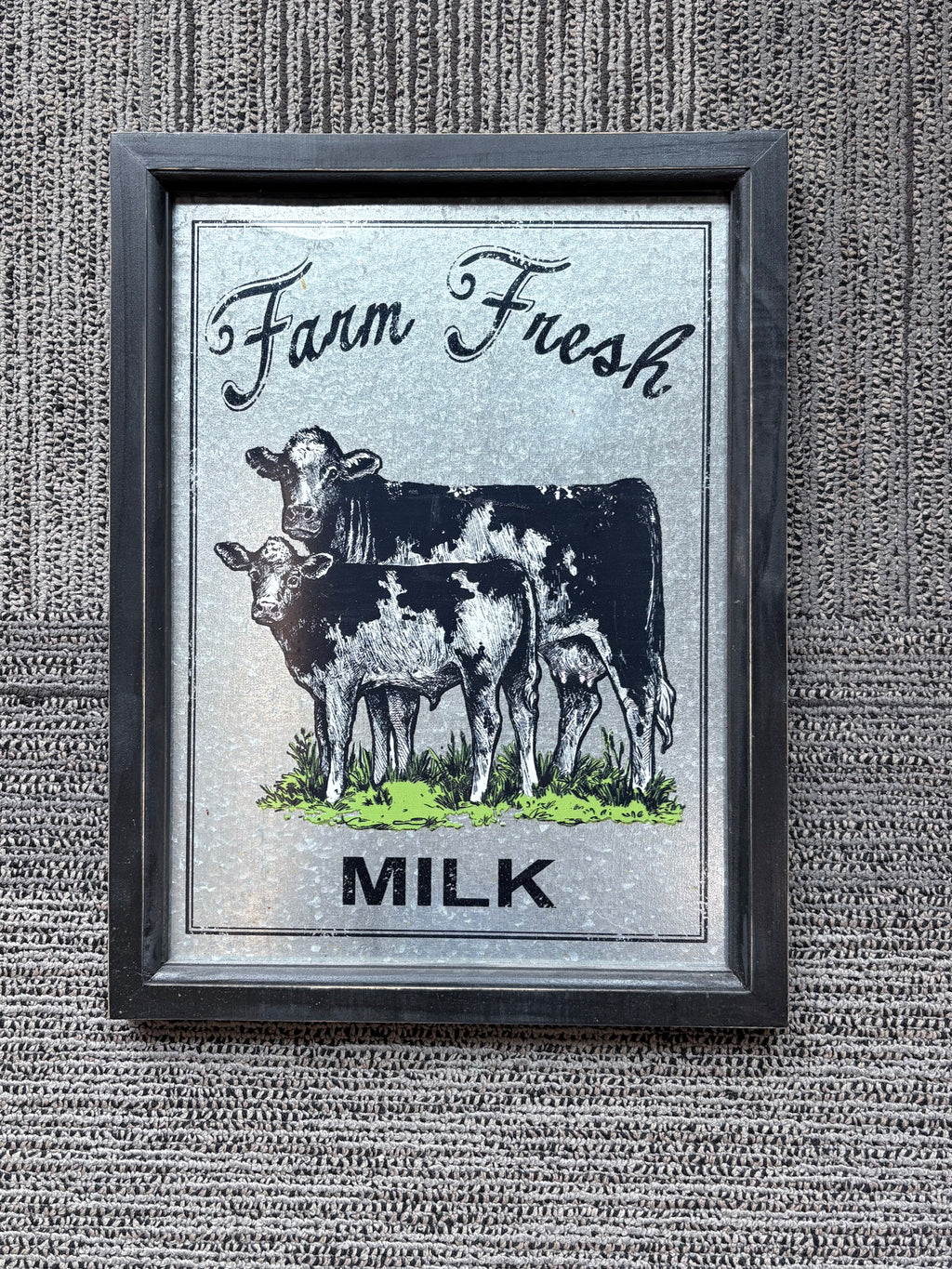 A framed vintage-style wall art print featuring two cows and the words 'Farm Fresh MILK' on a textured light background, mounted against a ribbed gray wall.