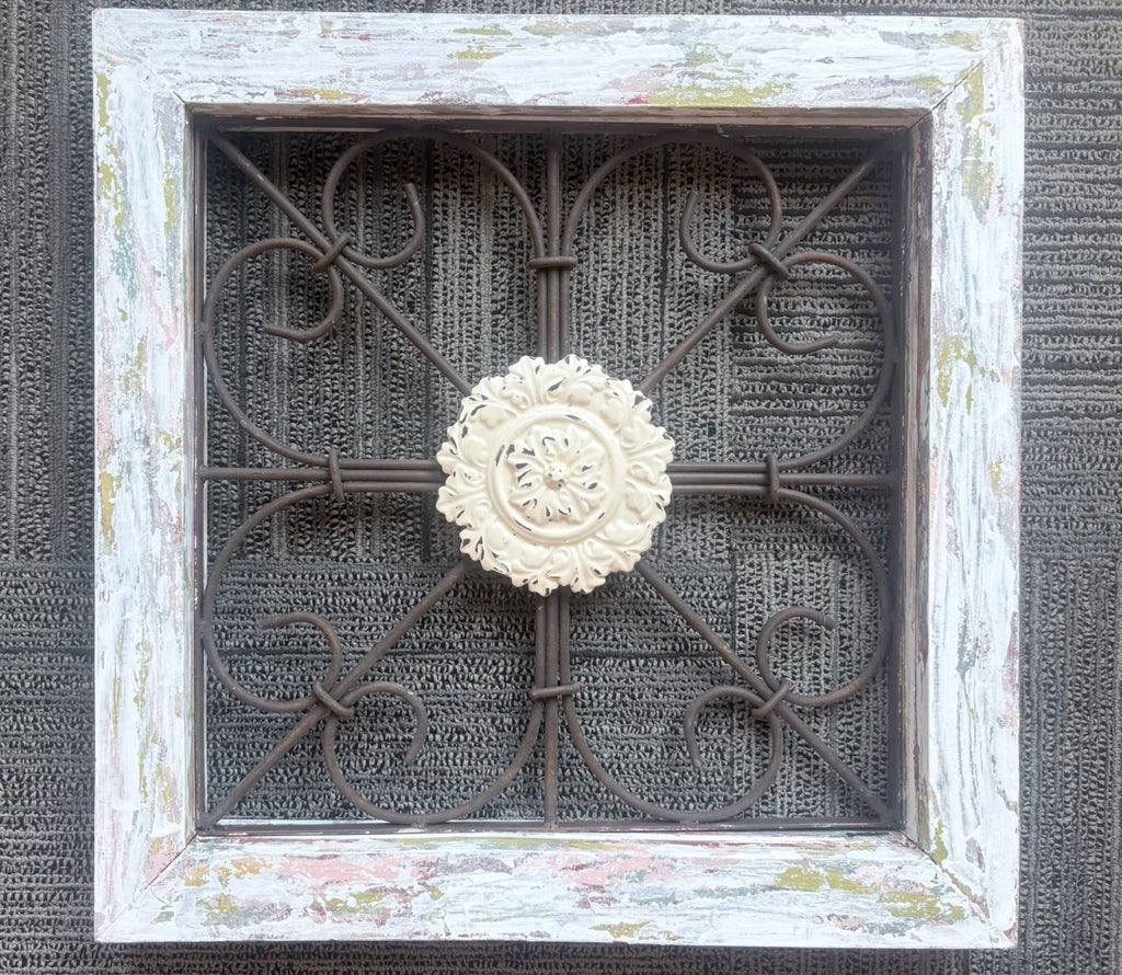 A square decorative wall art piece featuring a distressed white wood frame with a dark brown metal scrollwork center and a cream-colored carved floral medallion in the middle, set against a dark textured background.