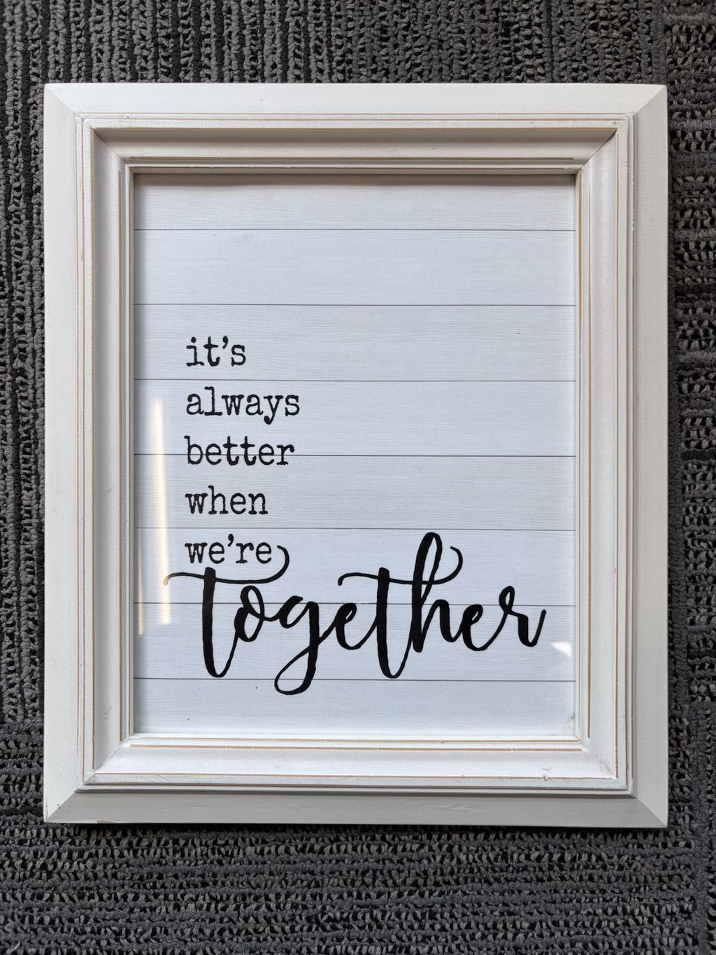 A framed typographic wall sign with the phrase “it’s always better when we’re together” in black text on a white lined-paper style background, set in a distressed off-white frame against a dark textured wall.