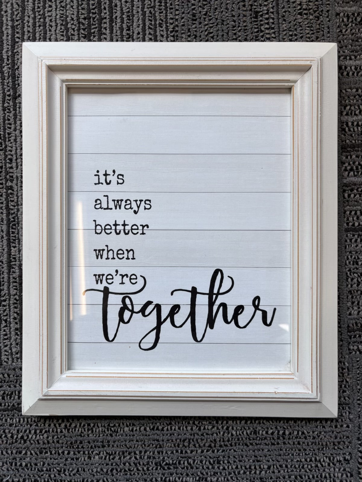 A framed typographic wall sign with the phrase “it’s always better when we’re together” in black text on a white lined-paper style background, set in a distressed off-white frame against a dark textured wall.
