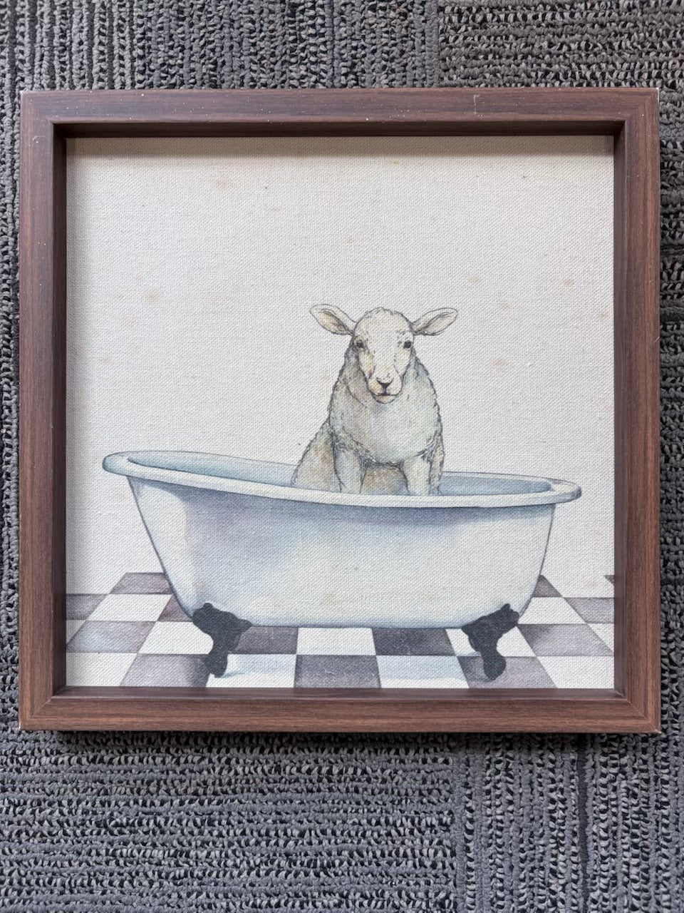 A framed art print featuring a watercolor-style illustration of a white lamb sitting inside a white clawfoot bathtub on a black-and-white checkered floor, set in a dark brown wooden frame against a dark textured background.