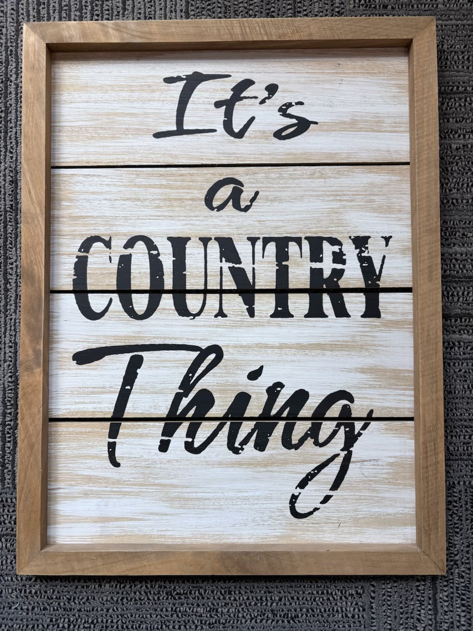 A framed rustic wooden sign with whitewashed planks and black lettering that reads 'It's a COUNTRY Thing' shown against a dark textured background.