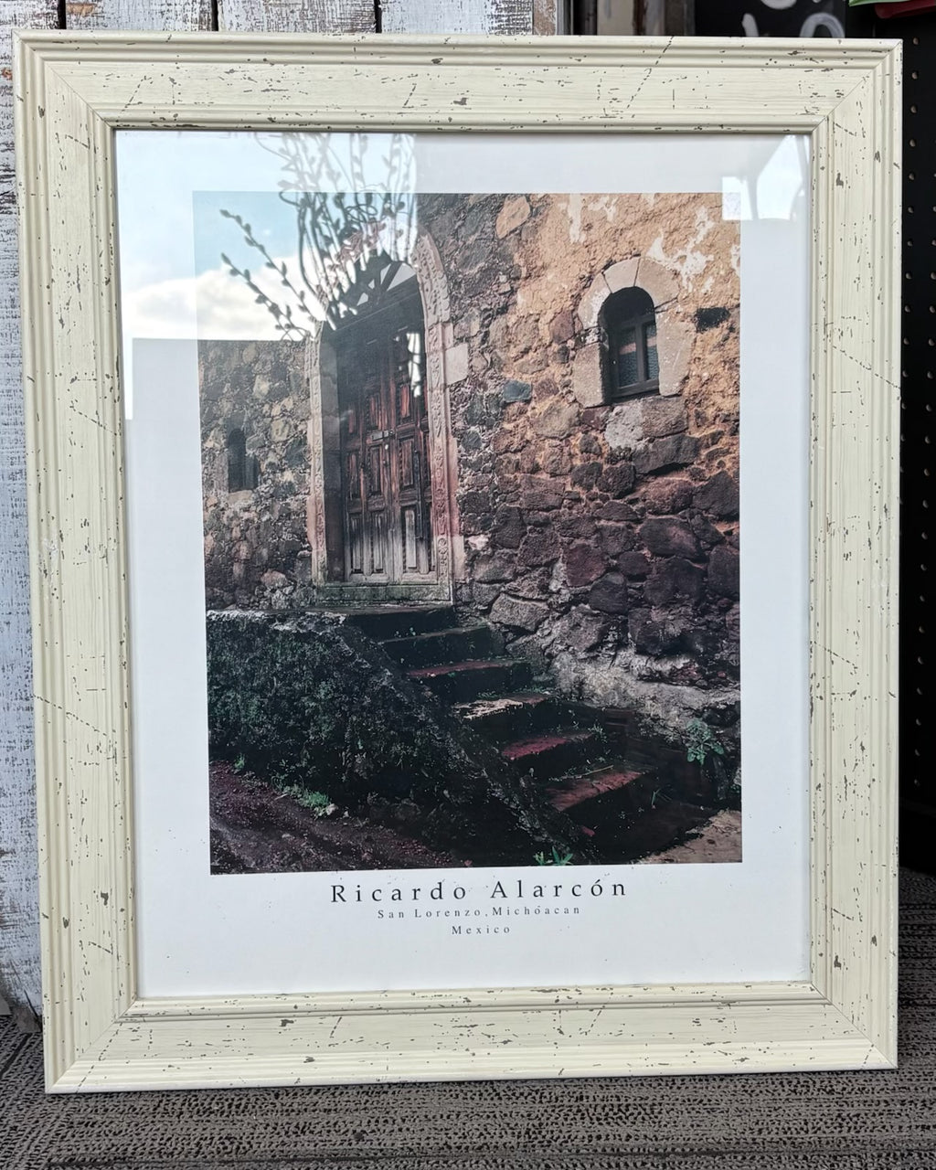 A framed art print featuring a photograph of a rustic stone building entrance with steps, signed 'Ricardo Alarcón' and displayed in a distressed off-white frame.