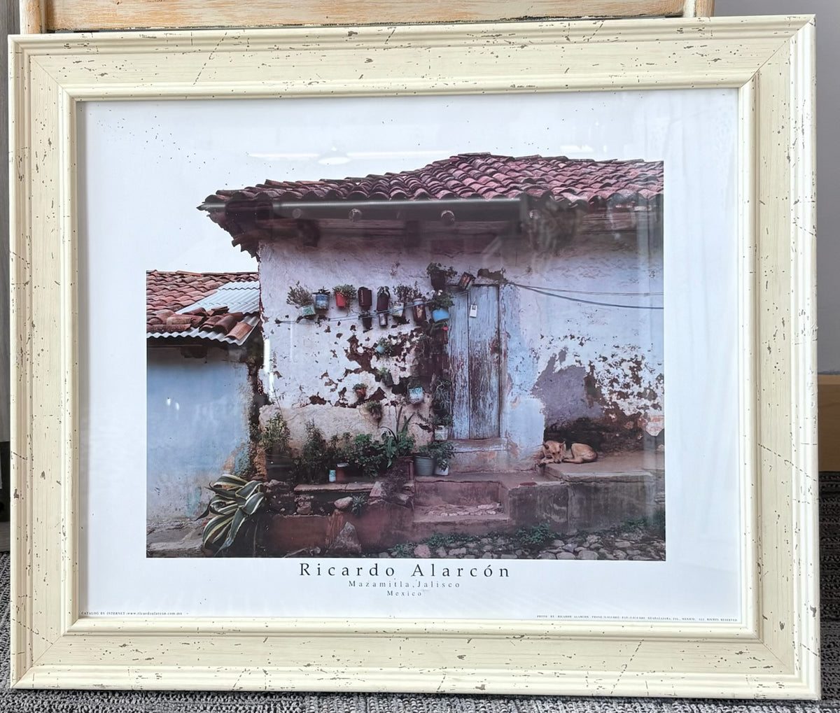 A framed art print by Ricardo Alarcón depicting a rustic white wall with a terracotta tiled roof, mounted in a distressed off-white wooden frame.