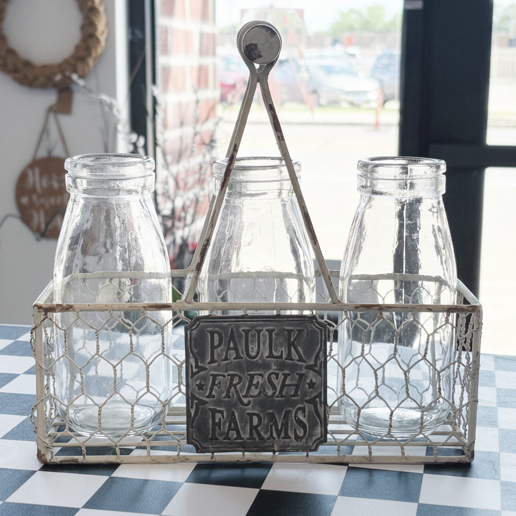 A rustic metal wire carrier basket holding three clear glass milk bottles and a dark metal sign that reads 'PAULK FRESH FARMS,' set on a checkered table in front of a window.