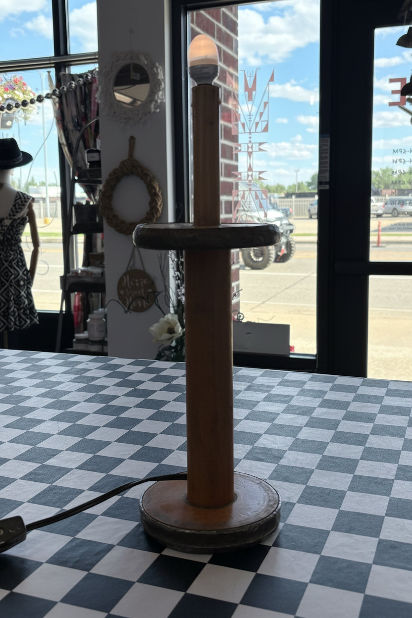 A rustic wooden table lamp shaped like a vintage rolling pin, featuring a cylindrical wooden handle, a horizontal wooden top, and a round base, set on a black and white checkered table in a shop interior.